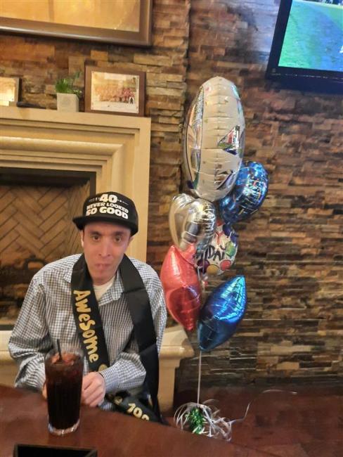 Adult man with a black baseball hat and striped shirt stands next to balloons to celebrate his birthday